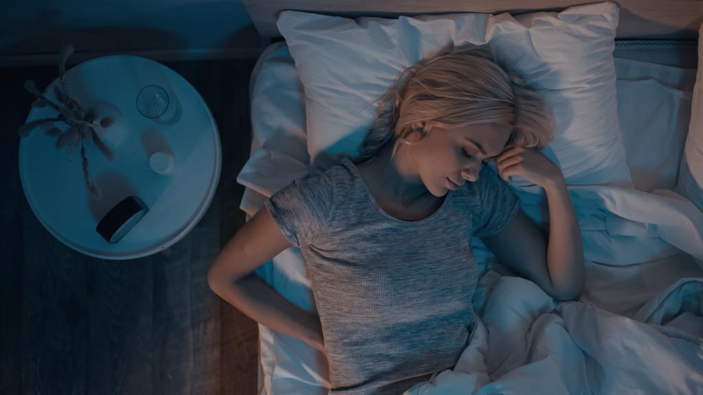 Bovenaanzicht van jonge vrouw slapen op bed in de buurt van pillen en glas water op nachtkastje  - Foto, afbeelding