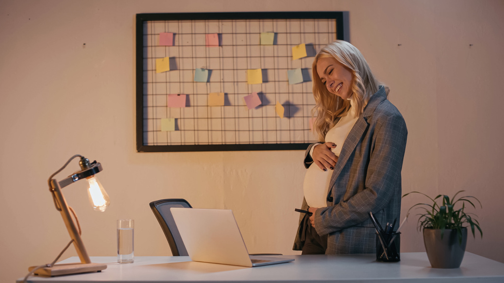 妊娠中のビジネスマンの笑顔夜にオフィスでラップトップでビデオ通話を持っている  - 写真・画像