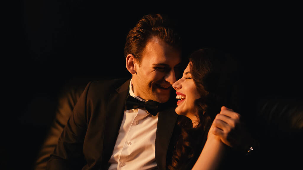 Lächelnder Mann im Anzug umarmt und flirtet mit Freundin isoliert auf schwarz  - Foto, Bild