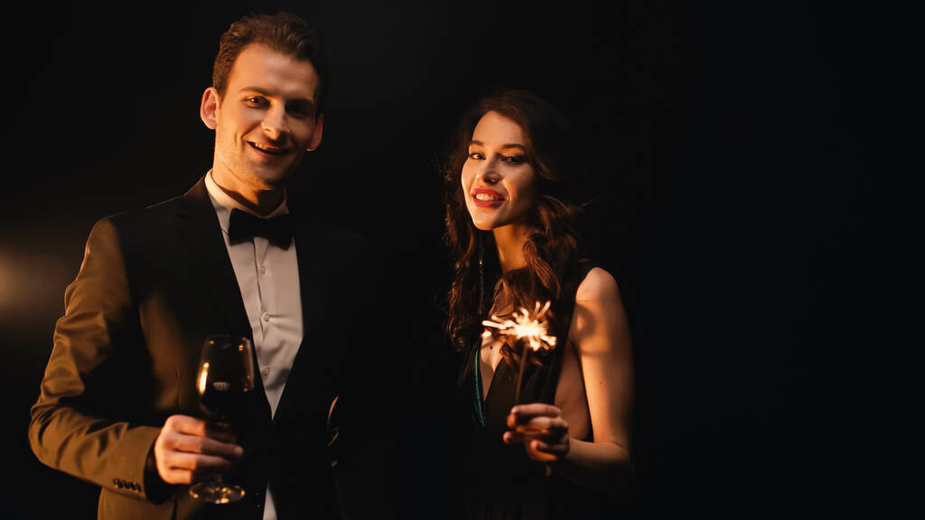 Uomo felice in possesso di un bicchiere di vino rosso e donna con sparkler guardando la fotocamera sul nero  - Foto, immagini
