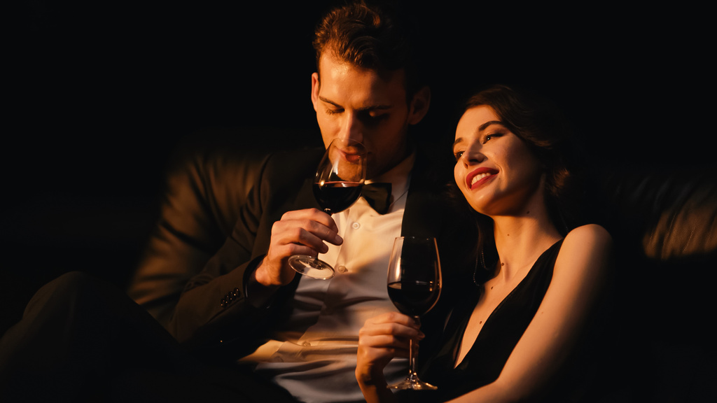 νεαρό και ευτυχισμένο ζευγάρι κρατώντας ποτήρια κόκκινο κρασί απομονωμένα σε μαύρο - Φωτογραφία, εικόνα