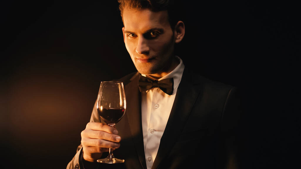 κομψό νεαρός άνδρας με κοστούμι με παπιγιόν κρατώντας ένα ποτήρι κρασί σε μαύρο  - Φωτογραφία, εικόνα
