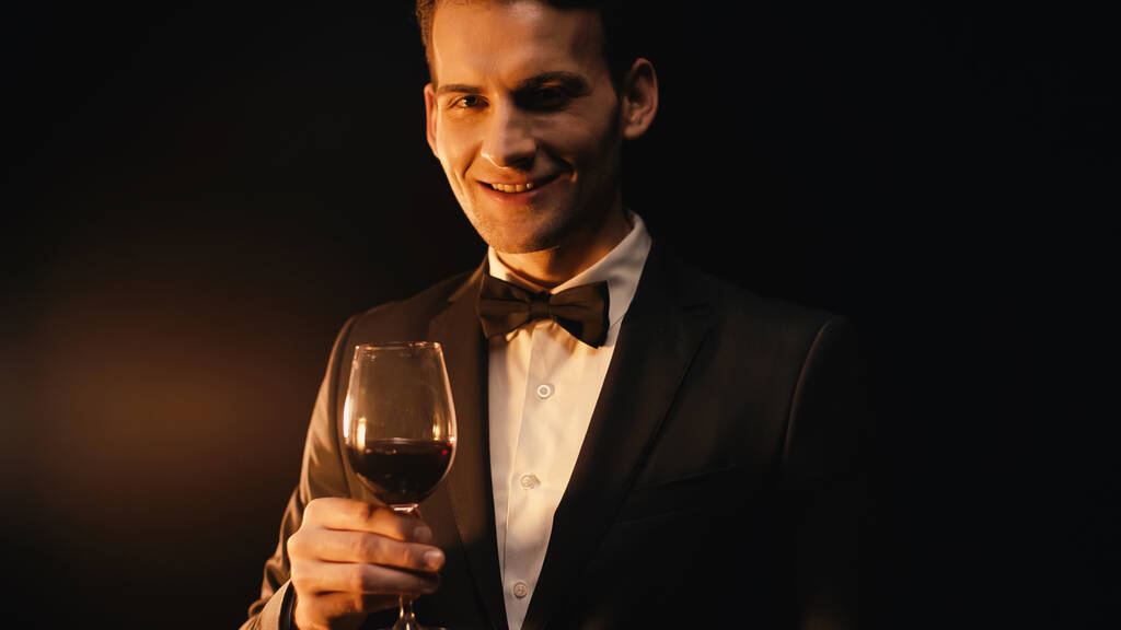 веселый молодой человек в костюме с галстуком-бабочкой держа бокал вина на черном  - Фото, изображение
