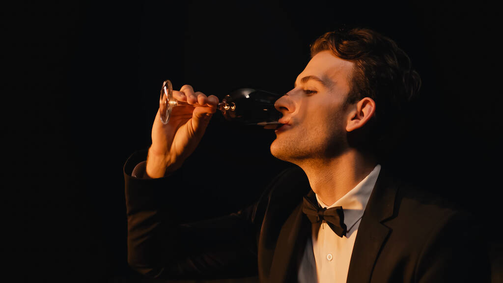 elegancki młody człowiek w garniturze z muszką trzymając szklankę i pijąc wino odizolowane na czarno  - Zdjęcie, obraz