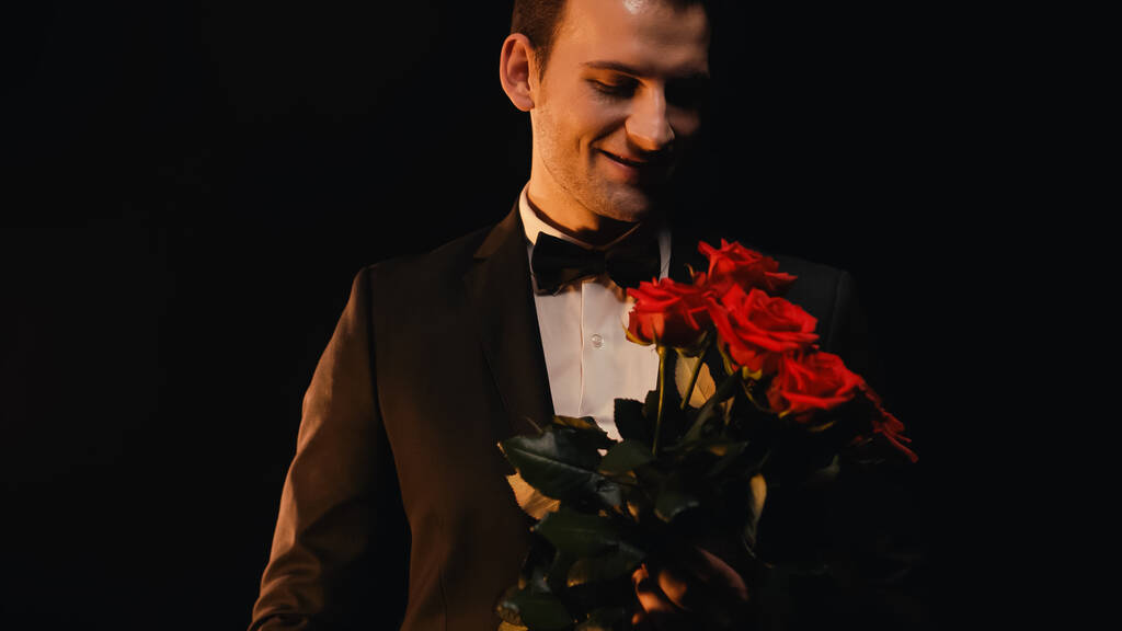 szczęśliwy młody człowiek patrząc na czerwone róże i uśmiechając się odizolowany na czarny  - Zdjęcie, obraz