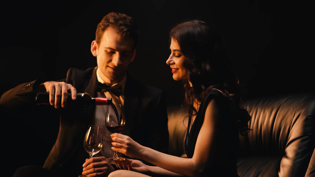 Счастливый мужчина наливает вино рядом с веселой женщиной держа стакан изолирован на черный - Фото, изображение