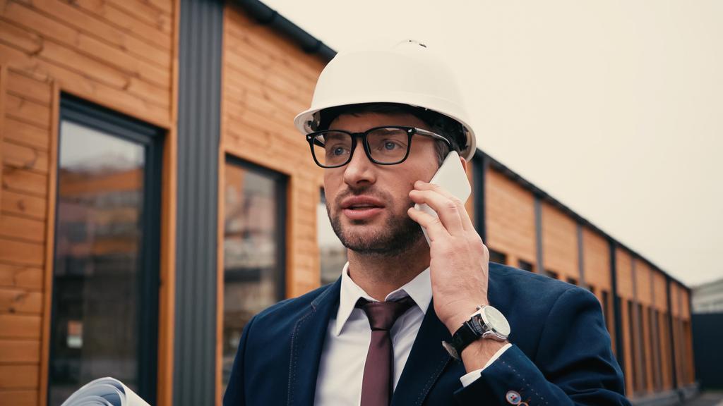Arkkitehdin kypärä puhuu älypuhelimella lähellä hämärtynyt rakennus ulkona  - Valokuva, kuva