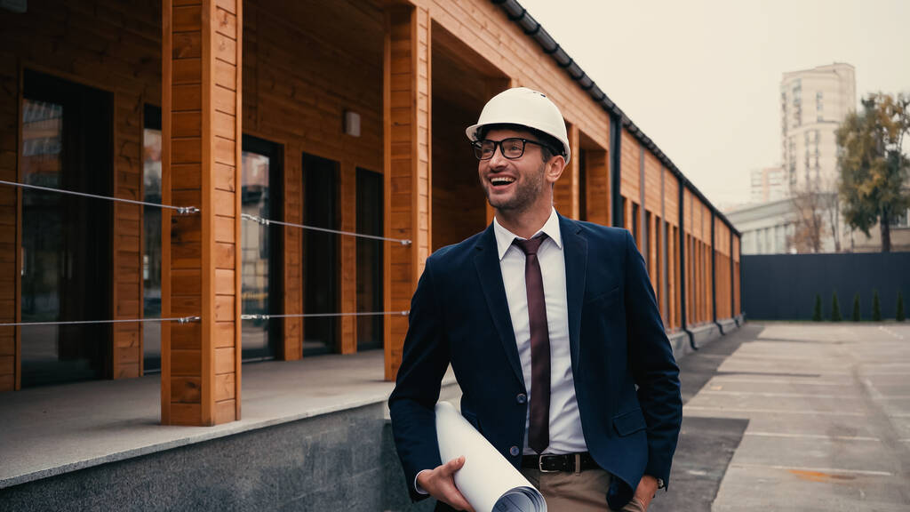Счастливый архитектор с чертежом, держащий руку в кармане рядом со зданием на открытом воздухе  - Фото, изображение