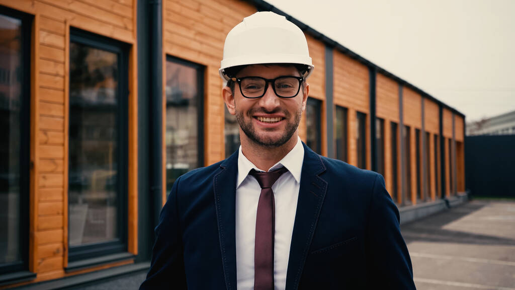 Lächelnder Ingenieur mit weißem Hut blickt in die Kamera in der Nähe von Gebäuden im Freien  - Foto, Bild