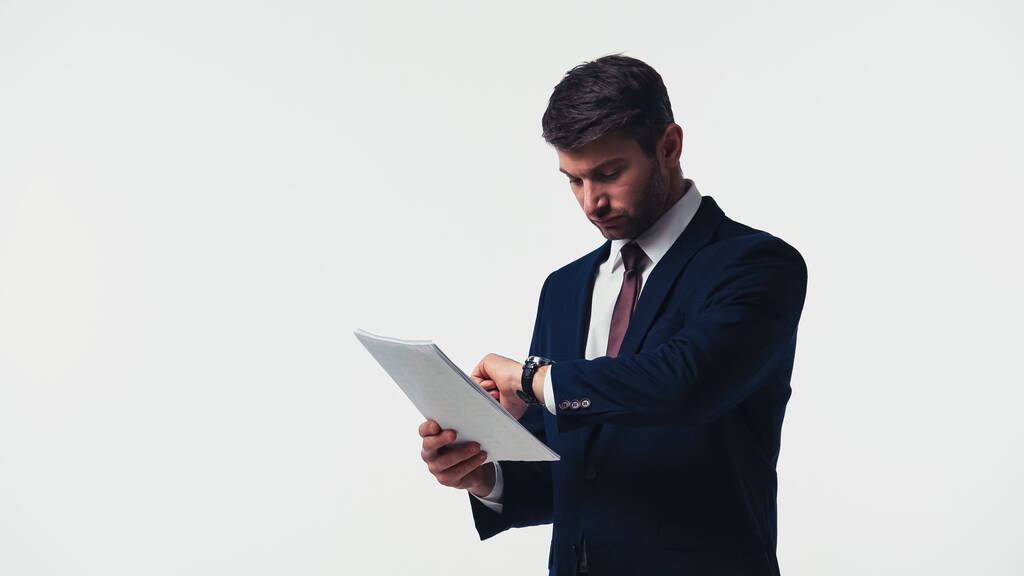 Бизнесмен смотрит на наручные часы, держа бумаги изолированными на белом - Фото, изображение
