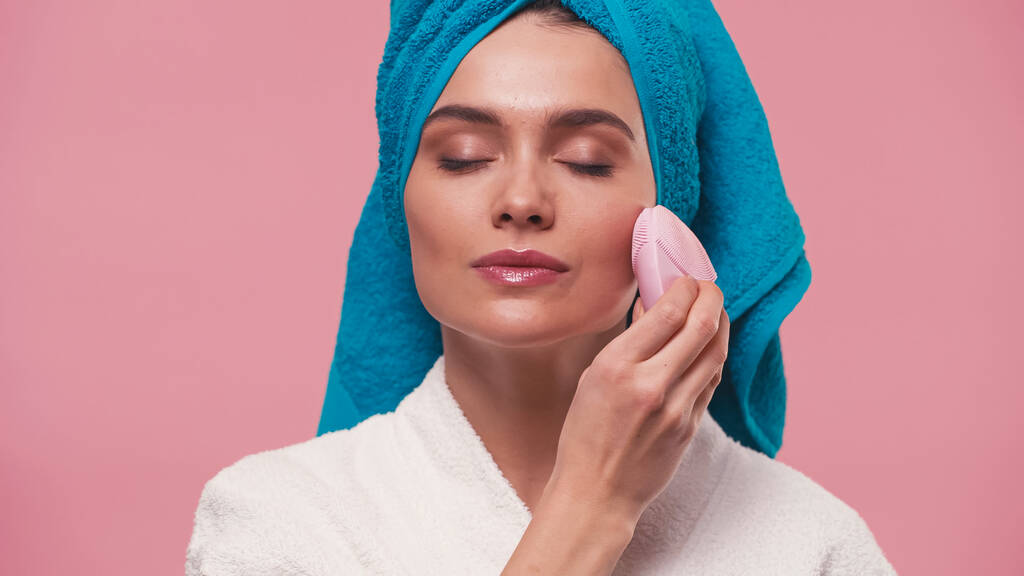young woman with closed eyes using silicone facial cleanser isolated on pink - Photo, Image