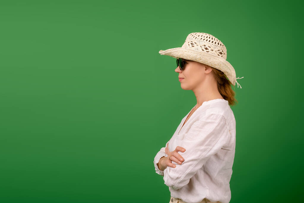 Ηρεμία αυτοπεποίθηση μεσήλικη γυναίκα σε ένα καπέλο, λευκό πουκάμισο και γυαλιά ηλίου σε ένα πράσινο φόντο. Στέκεται πλάγια. Θέση για το κείμενο σας. - Φωτογραφία, εικόνα