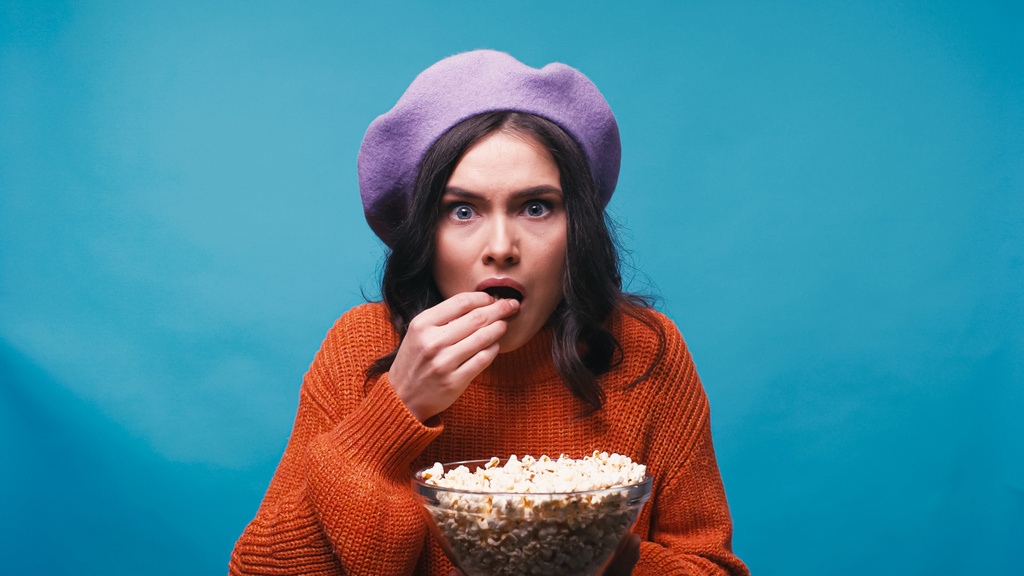 mujer preocupada en boina púrpura viendo película y comiendo palomitas aisladas en azul - Foto, imagen