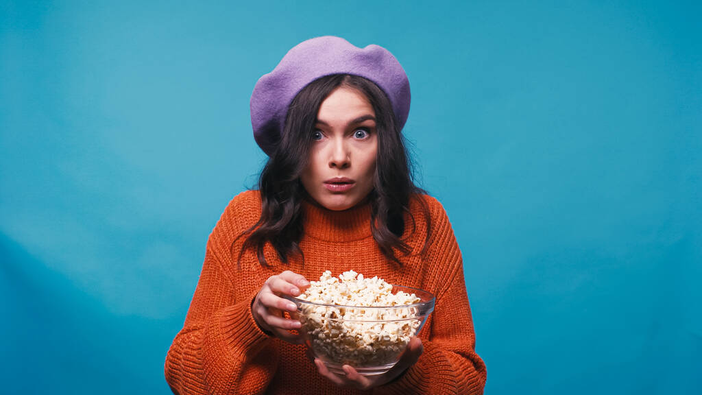 vzrušená žena drží misku s popcornem při sledování filmu izolované na modré - Fotografie, Obrázek