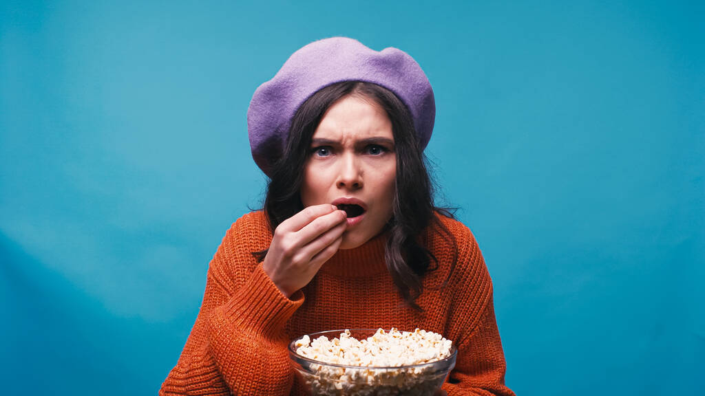 nervous woman eating popcorn while watching exciting film isolated on blue - Photo, Image