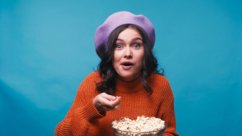 mujer sorprendida en jersey y boina viendo emocionante película y comiendo palomitas de maíz aisladas en azul - Foto, imagen