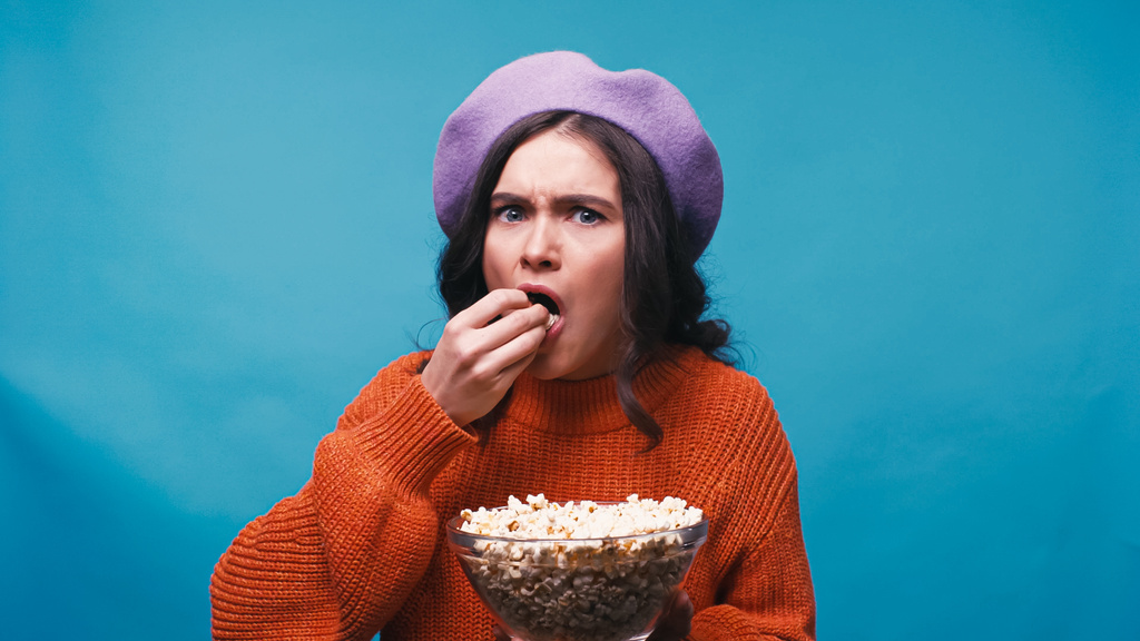 mujer preocupada en boina lila comiendo palomitas de maíz mientras ve la película aislada en azul - Foto, Imagen