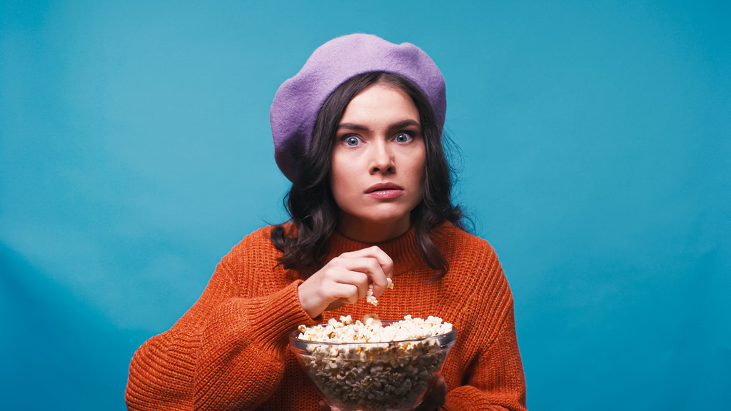 schockierte Frau hält Popcorn in der Hand, während sie Film isoliert auf blauem Grund anschaut - Foto, Bild