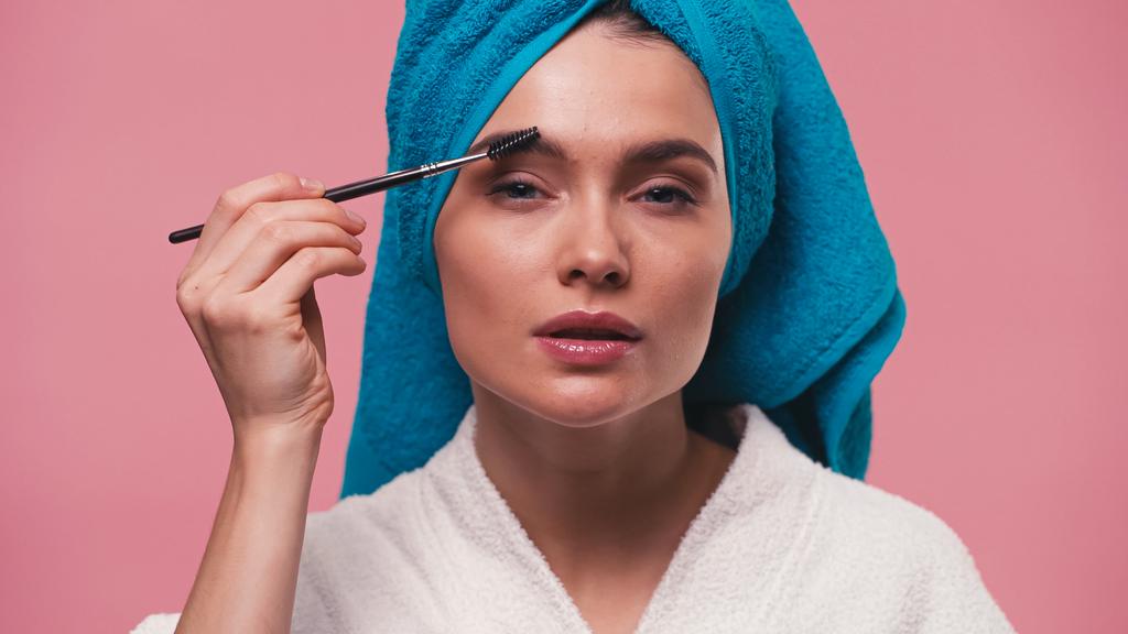 woman with towel on head styling eyebrows with eyebrow brush isolated on pink - Photo, Image