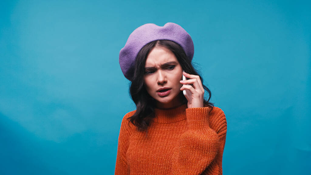 τεταμένη γυναίκα σε μωβ μπερέ μιλώντας στο κινητό τηλέφωνο απομονωμένη σε μπλε - Φωτογραφία, εικόνα