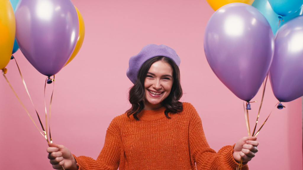 fröhliche Frau in Baskenmütze und Pullover mit bunten Luftballons auf rosa - Foto, Bild