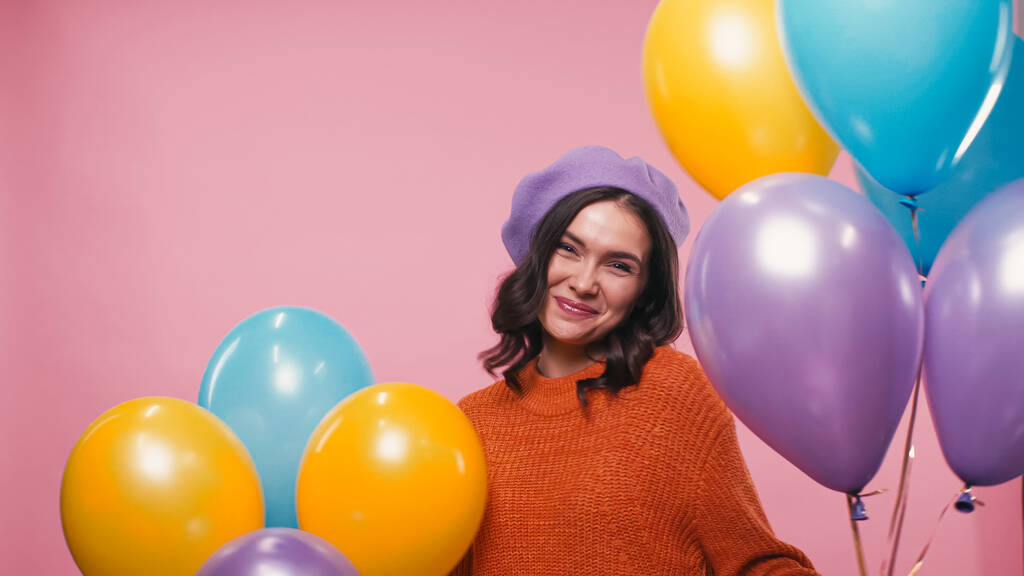 ευτυχισμένη γυναίκα σε μπερέ και πουλόβερ χαμογελώντας κοντά εορταστικά μπαλόνια που απομονώνονται σε ροζ - Φωτογραφία, εικόνα