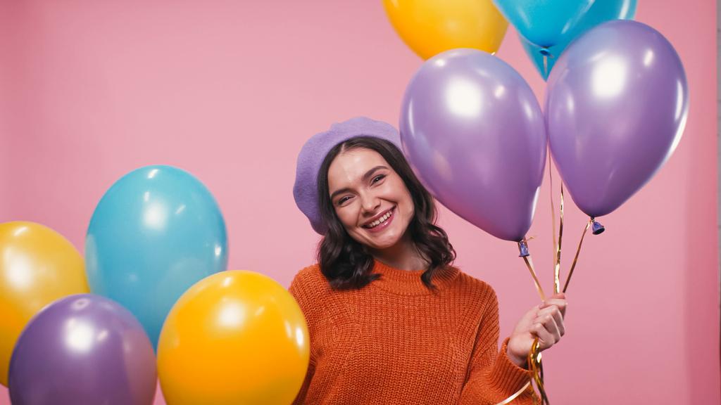 wesoła kobieta w ciepłym swetrze i berecie uśmiechnięta do kamery w pobliżu kolorowych balonów odizolowanych na różowo - Zdjęcie, obraz