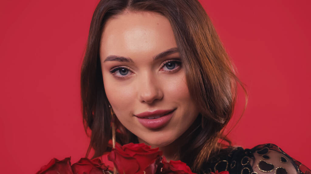 bonita joven sonriendo cerca de rosas aisladas en rojo - Foto, imagen