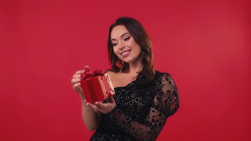 feliz joven mujer sonriendo y mirando caja de regalo aislado en rojo - Foto, imagen