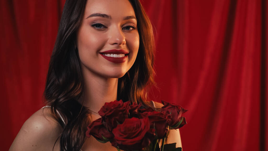 Ευτυχισμένη νεαρή γυναίκα με φωτεινά χείλη χαμογελώντας κοντά σε τριαντάφυλλα στο κόκκινο - Φωτογραφία, εικόνα