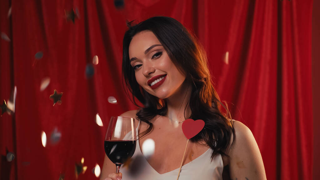 happy woman holding glass of wine near falling confetti on red - Photo, Image