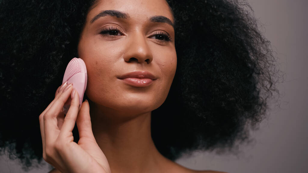 ricci donna afroamericana utilizzando dispositivo di pulizia del silicone sul viso isolato su grigio  - Foto, immagini