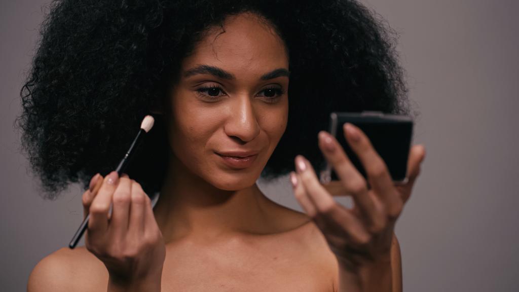 joven afroamericana mujer con cepillo cosmético mirando espejo aislado en gris - Foto, Imagen