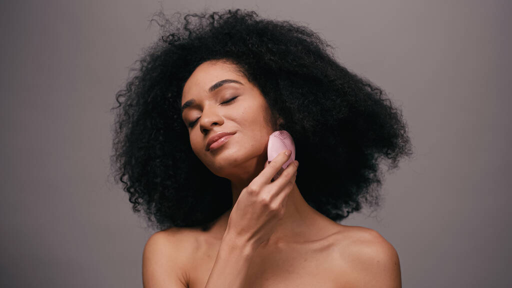 piacere afroamericano donna con spalle nude utilizzando dispositivo di pulizia del silicone sul viso isolato su grigio  - Foto, immagini