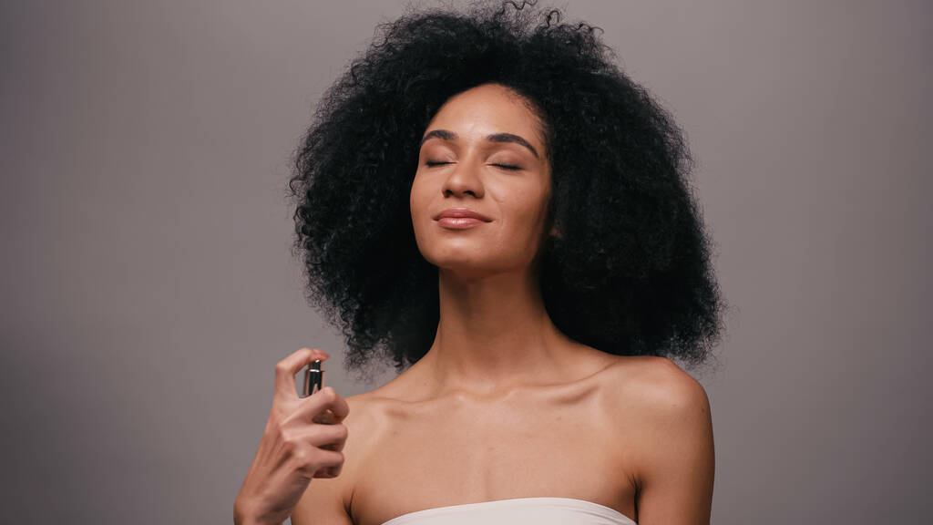 sorrindo afro-americano mulher pulverizando perfume isolado em cinza - Foto, Imagem