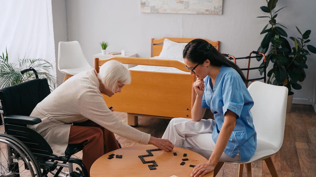 vanhempi nainen pyörätuolissa pelaa dominoa nuoren sairaanhoitajan kanssa - Valokuva, kuva