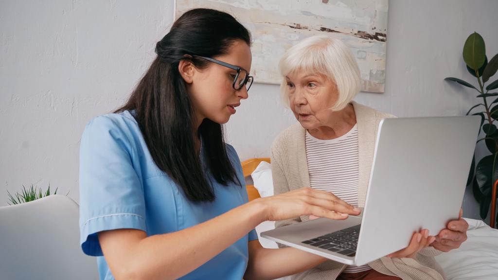 brünette geriatrische Krankenschwester zeigt Seniorin, wie man Laptop benutzt - Foto, Bild