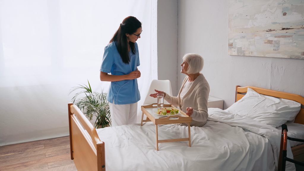 μελαχρινή νοσοκόμα μιλάει με χαρούμενη ηλικιωμένη ασθενή κοντά στο δίσκο πρωινού στο κρεβάτι  - Φωτογραφία, εικόνα