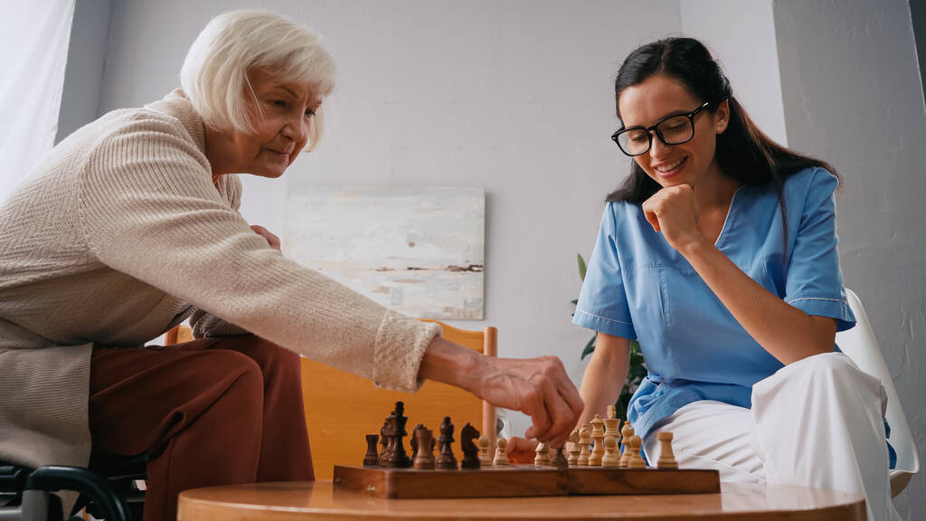 ηλικιωμένη γυναίκα και χαρούμενη νοσοκόμα παίζουν σκάκι στο γηροκομείο - Φωτογραφία, εικόνα