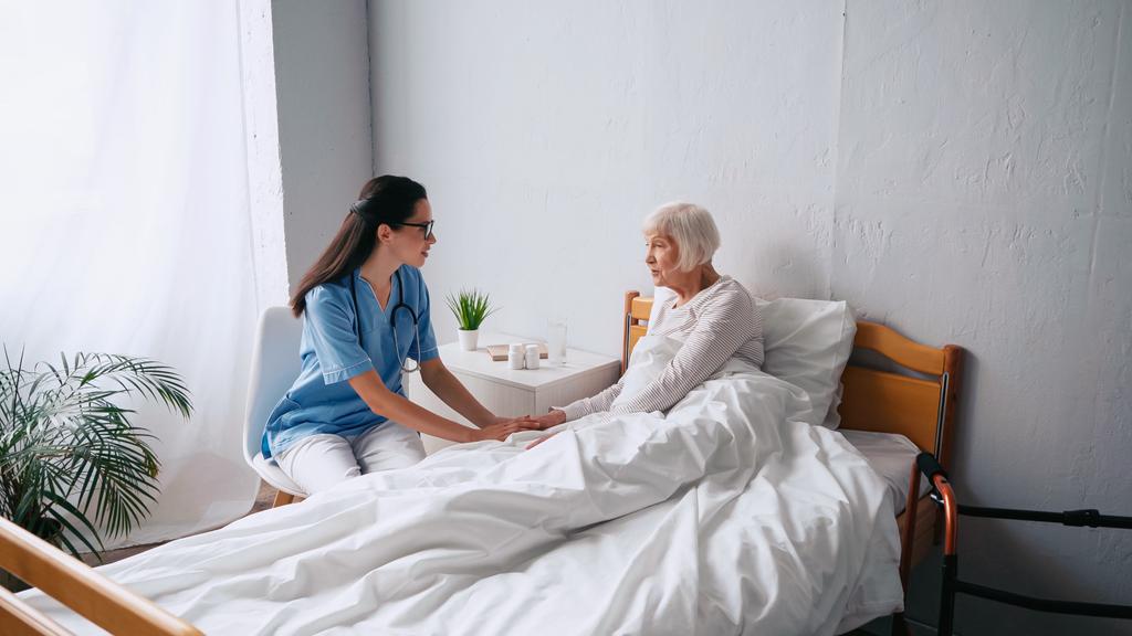 enfermeira morena em óculos e mulher idosa com cabelos grisalhos falando na clínica - Foto, Imagem