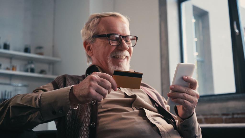 веселий старший чоловік з кредитною карткою тримає мобільний телефон під час покупок онлайн вдома
 - Фото, зображення