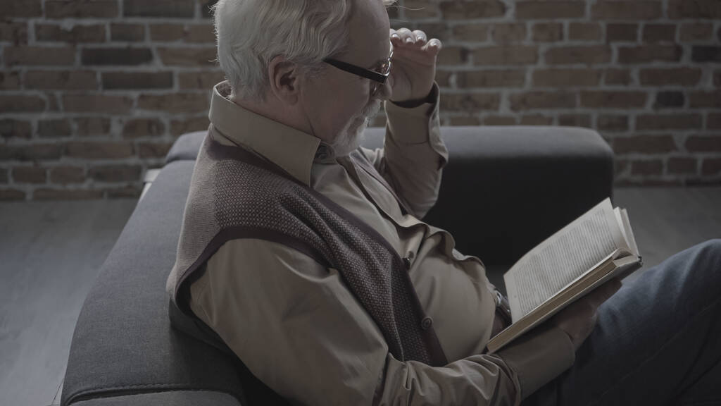 Seitenansicht eines älteren Mannes mit Brille, der zu Hause Buch liest  - Foto, Bild