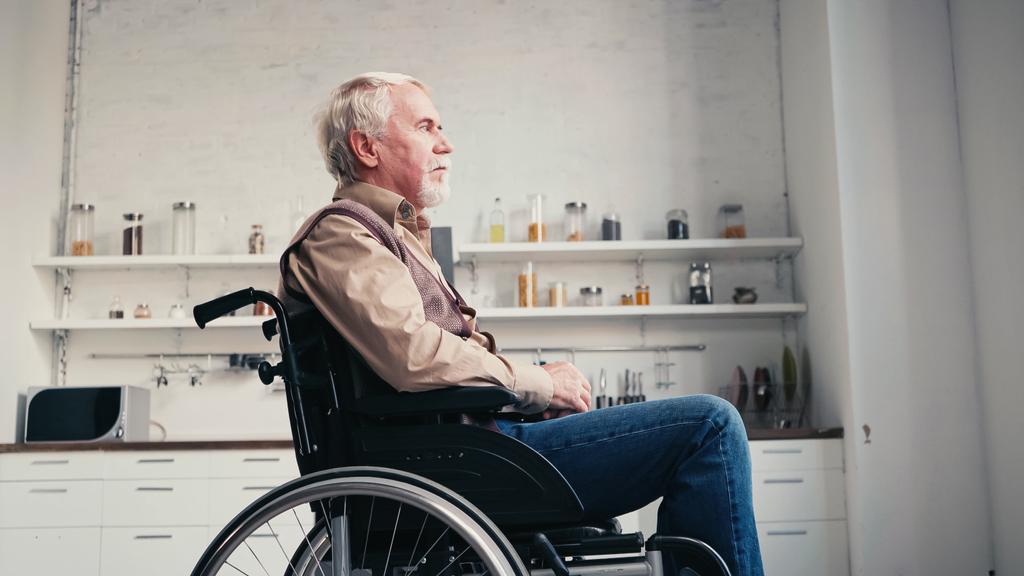 Seitenansicht eines bärtigen Rentners im Rollstuhl, der zu Hause wegschaut  - Foto, Bild