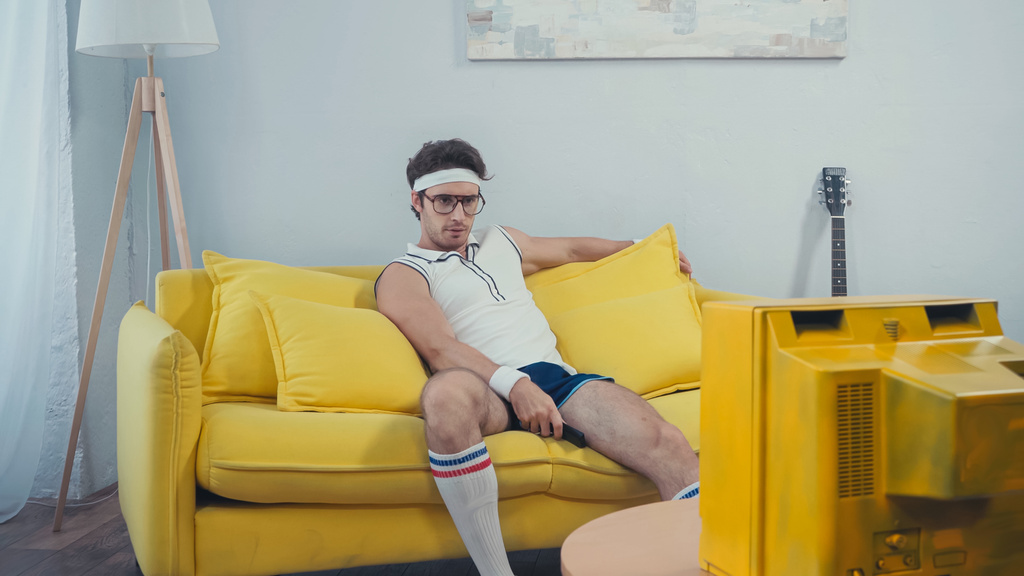 lazy man in sportswear watching retro tv sitting on sofa in living room - Photo, Image