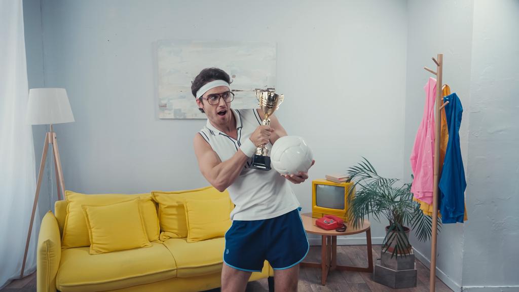 desportista animado segurando copo de troféu e bola de esportes enquanto está em casa - Foto, Imagem