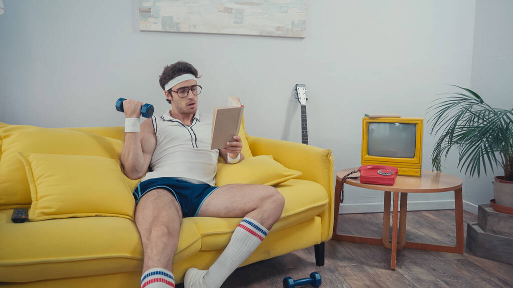Sportler hebt Hantel beim Lesen auf Sofa im Wohnzimmer - Foto, Bild