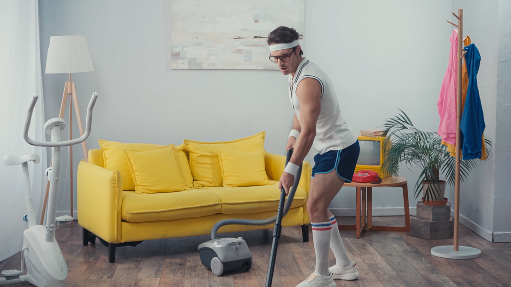 молодий чоловік у спортивному одязі вакуумна прибиральниця вітальня, концепція ретро спорту
 - Фото, зображення