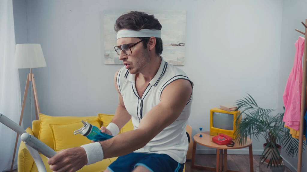 sportsman working out on exercise bike and holding sports bottle in living room, retro sport concept - Photo, Image