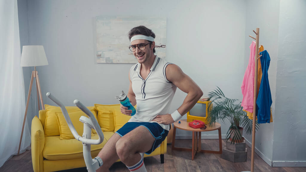šťastný sportovec školení na cvičení na kole a držení sportovní láhev v obývacím pokoji, retro sportovní koncept - Fotografie, Obrázek