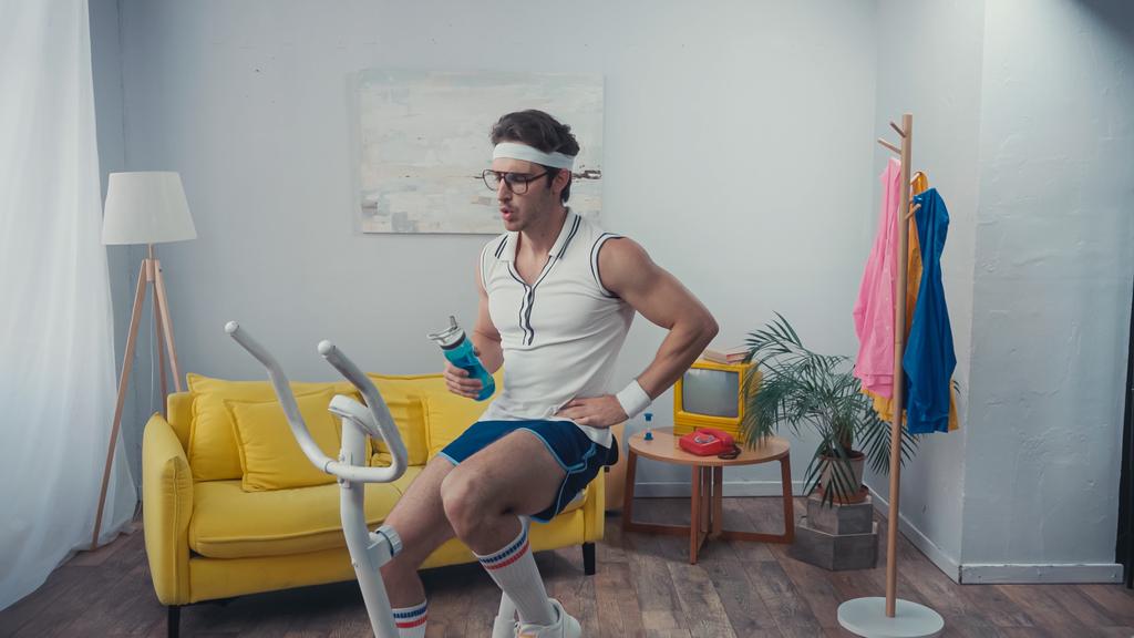 entraînement sportif fatigué sur vélo d'appartement et tenue de bouteille de sport dans le salon, concept sportif rétro - Photo, image